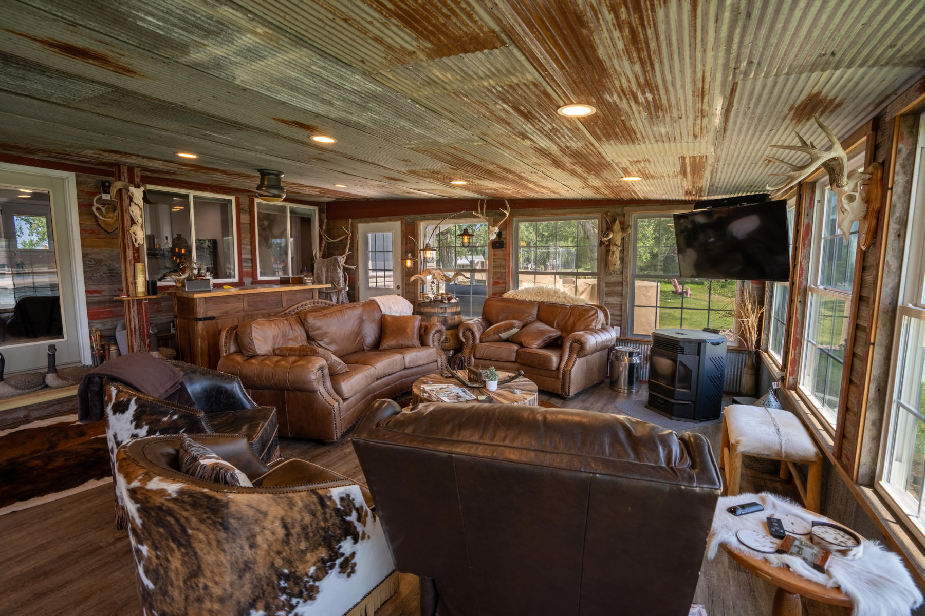 nebraska-waterfowl-hunting-lodge3 - Xtreme Waterfowlers - Nebraska Guided Duck & Goose Hunts Rustic living room with brown leather sofas, cowhide chairs, antler decor, a corrugated metal ceiling, and a wall-mounted TV by large windows.